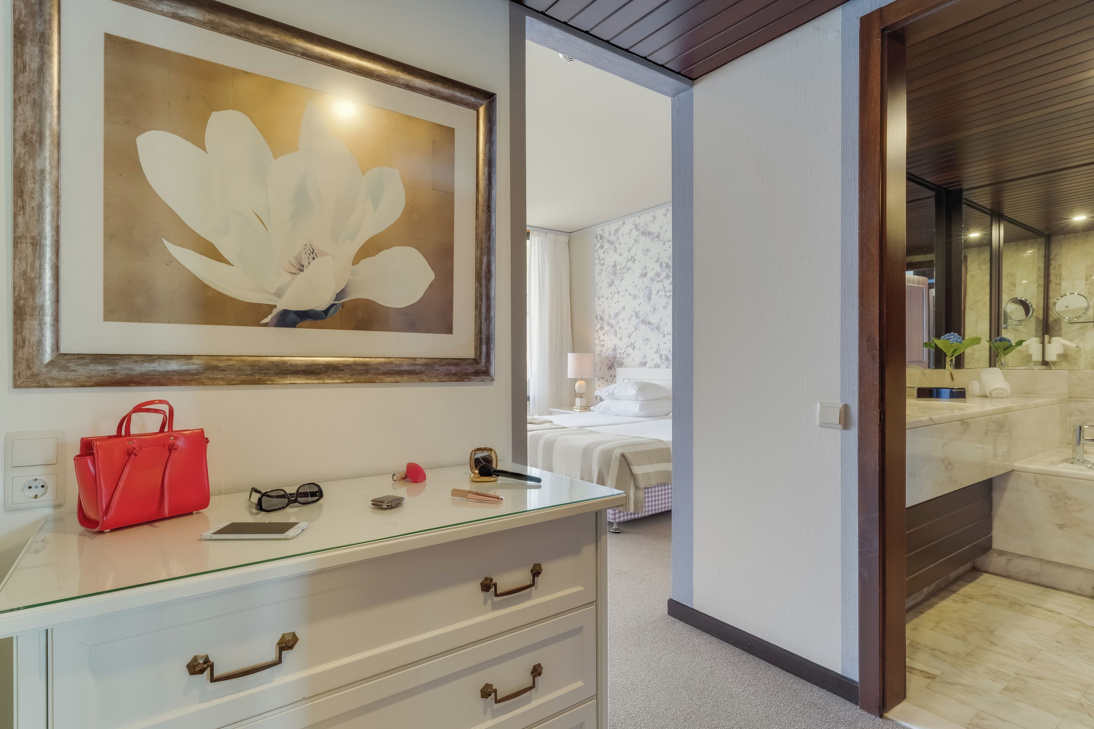 Quarto de hotel com cômoda de vidro, obra de arte floral emoldurada, bolsa vermelha e cama com colcha quadriculada branca e preta.