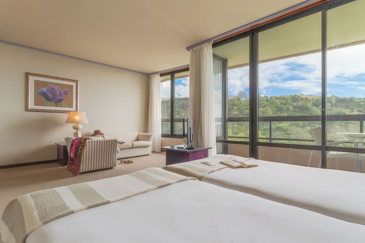 Ett hotellrum med två sängar, en soffa, ett skrivbord, en lampa och ett stort fönster med utsikt över grönområden.