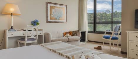 A modern hotel room with a bed, a desk, a chair, a sofa, and a large window offering a view of the outdoors.