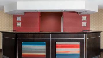 Um balcão de recepção de hotel moderno, com esquema de cores vermelho e preto, dois quadros emoldurados e uma placa indicando a presença de um gerente.
