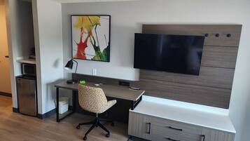 A modern bedroom with a bed, a desk, a chair, a TV, and a small kitchenette.