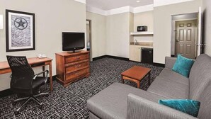 A hotel room with a gray sofa, a wooden desk with a TV, a black office chair, a patterned carpet, a small wooden table, and a kitchenette with a microwave. - Quality Inn and Suites (Rosenberg)