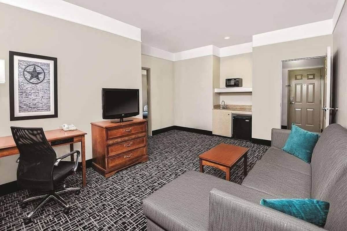 A hotel room with a gray sofa, a wooden desk with a TV, a black office chair, a patterned carpet, a small wooden table, and a kitchenette with a microwave.