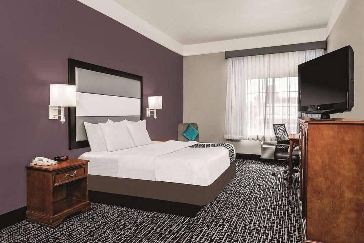 A hotel room with a large bed, a desk with a flat-screen TV, a nightstand, and a window with curtains.
