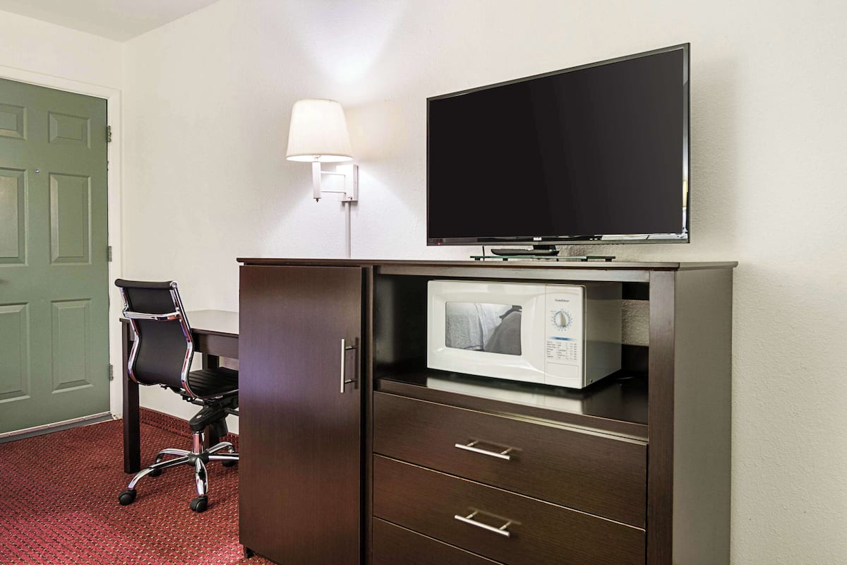 Quarto de hotel com televisão de tela plana presa acima de um armário de madeira, micro-ondas dentro do armário, cadeira de escritório preta e luminária na parede.