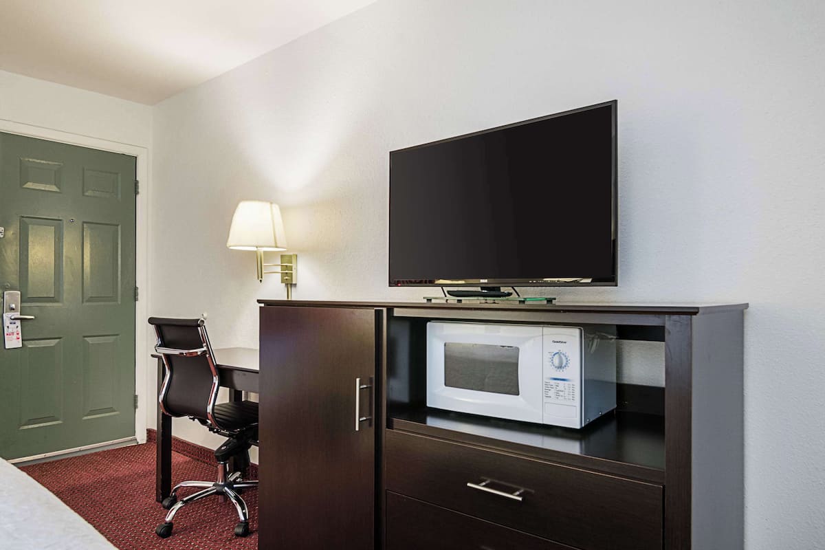 Quarto de hotel com cama, escrivaninha com cadeira, televisão, luminária e micro-ondas.