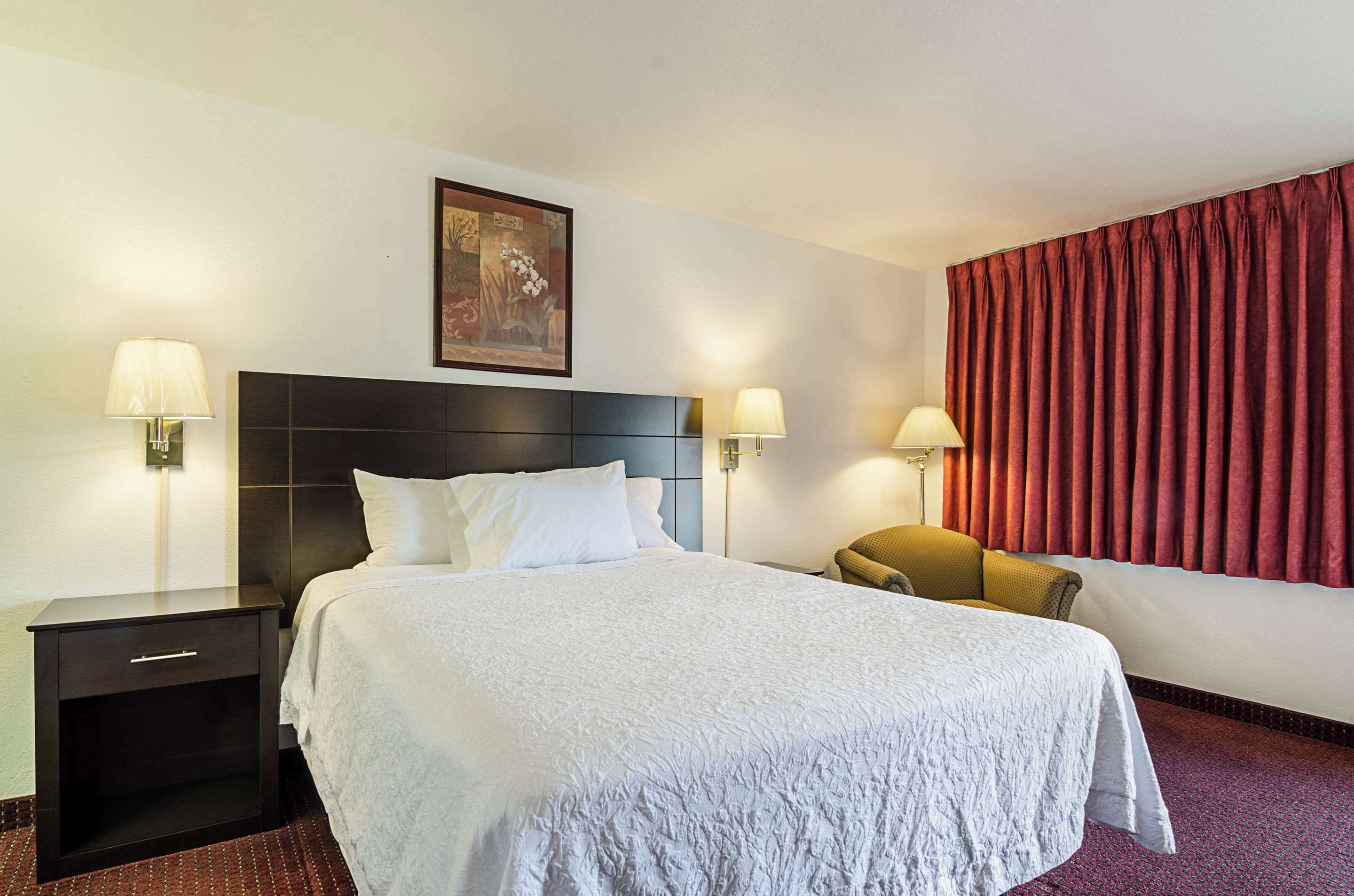 A hotel room with a large bed, two bedside tables with lamps, a chair, a painting on the wall, and red curtains.