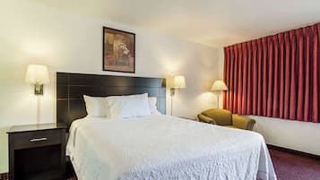 A hotel room with a large bed, two bedside tables with lamps, a chair, a painting on the wall, and red curtains.