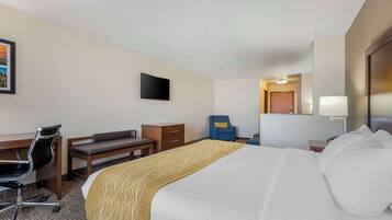 Habitación de hotel con una cama grande, un escritorio con silla, una mesita pequeña, un sillón azul, un televisor montado en la pared y una lámpara de noche.