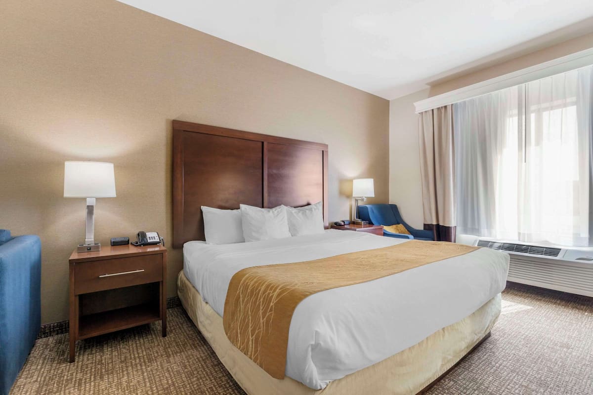Ein Hotelzimmer mit einem großen Bett, einem Holzkopfstück, zwei Nachttischen mit Lampen, einem blauen Sessel und einem großen Fenster mit Vorhängen.