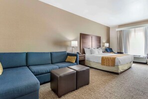 A hotel room with a bed, a blue sofa, two ottomans, a nightstand, a lamp, and a window with curtains.