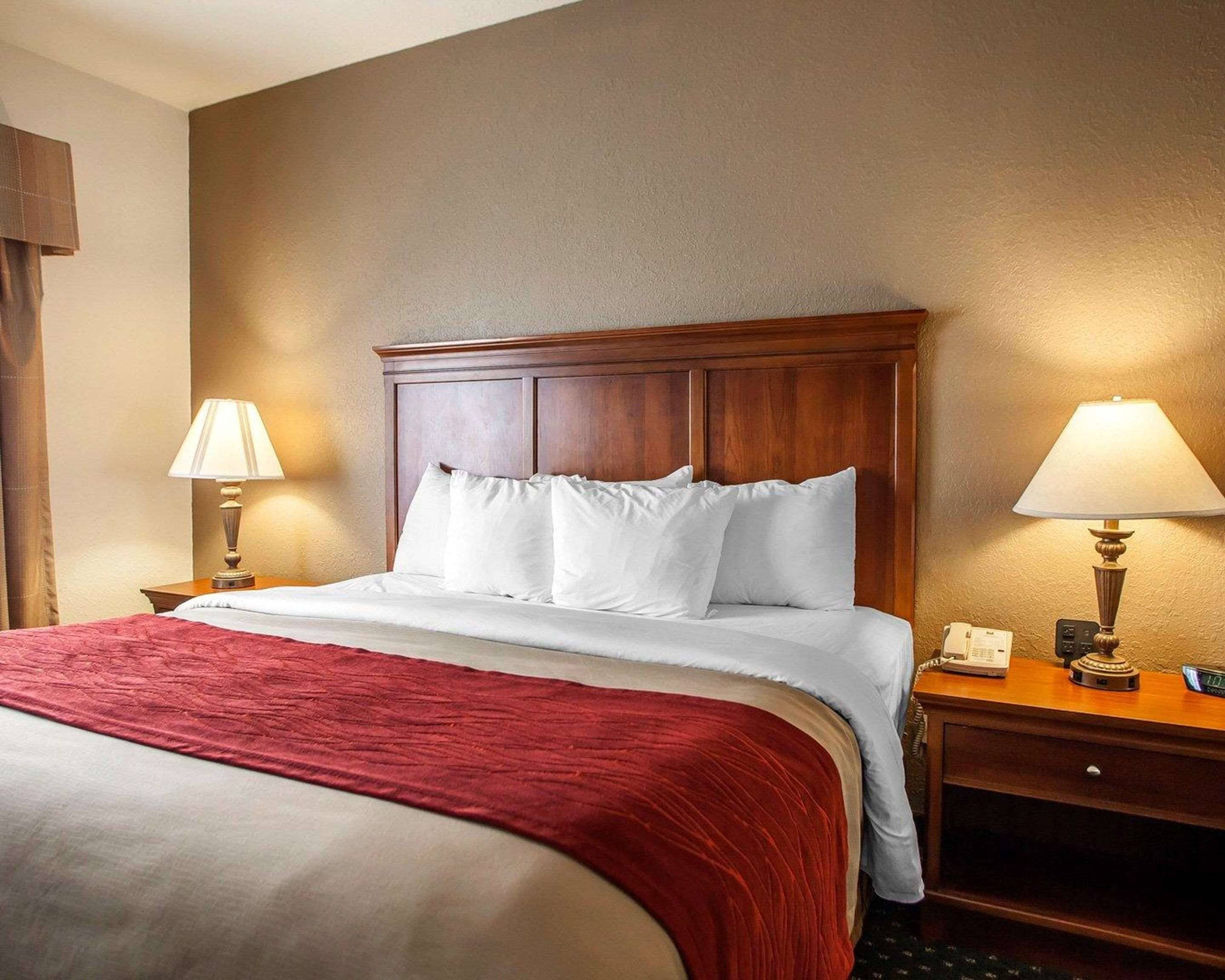 Une chambre d’hôtel avec une tête de lit en bois, une couvre-lit rouge, deux lampes de chevet et une table de chevet avec un téléphone et une horloge numérique.