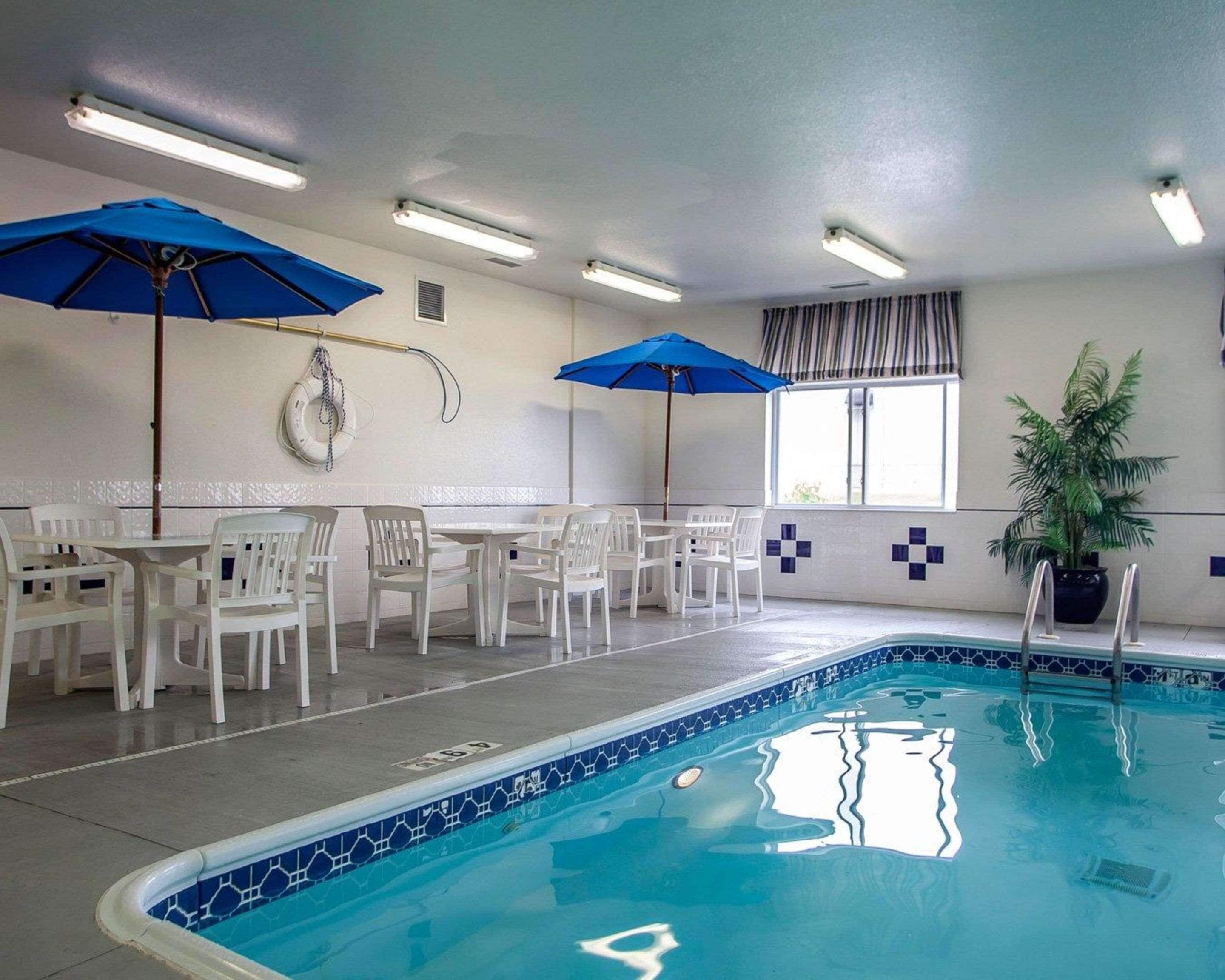 Zona de piscina con sillas blancas y sombrillas azules, una planta en maceta y un ventanal con cortinas.