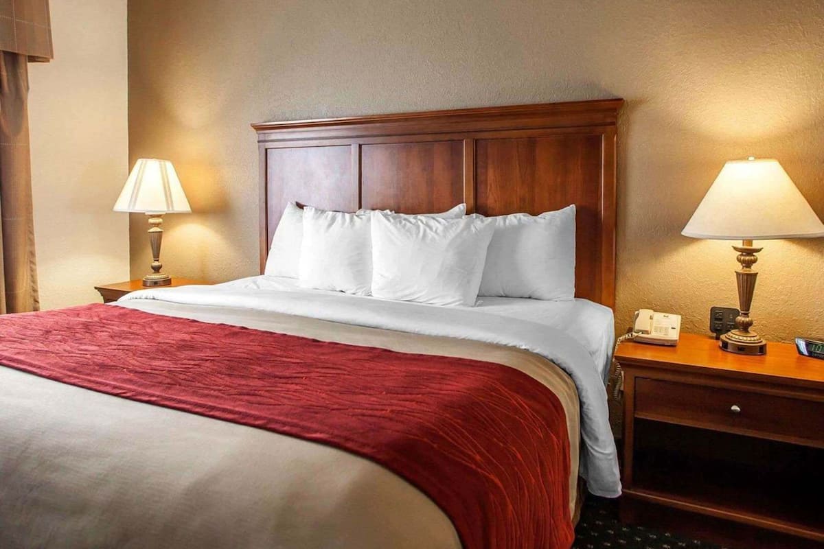 Habitación de hotel con cabecera de madera, colcha roja, dos lámparas de noche, mesita de noche con teléfono y reloj, y panel de control en la pared.