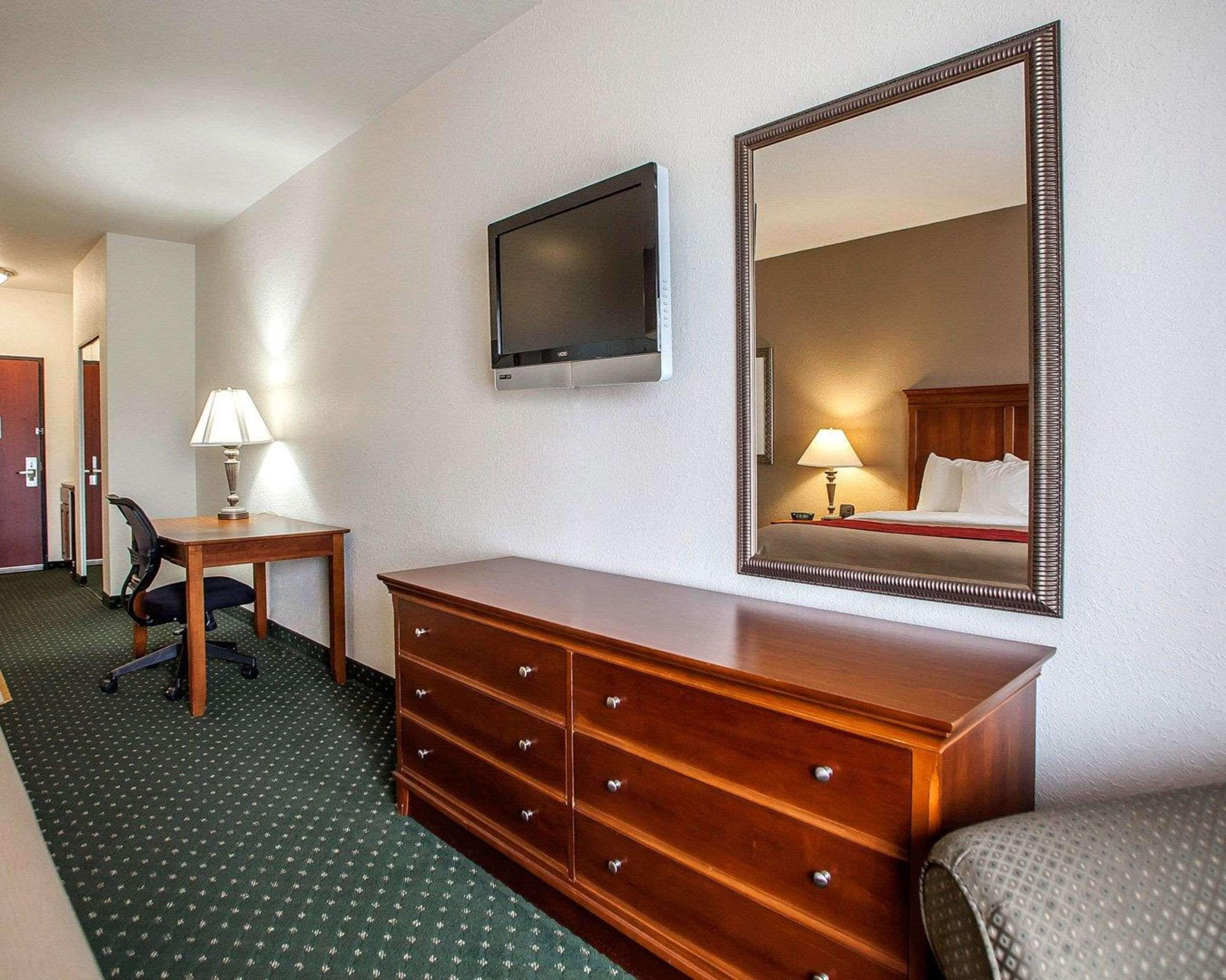 A hotel room with a wooden dresser, a desk with a lamp, a television, and a large mirror.