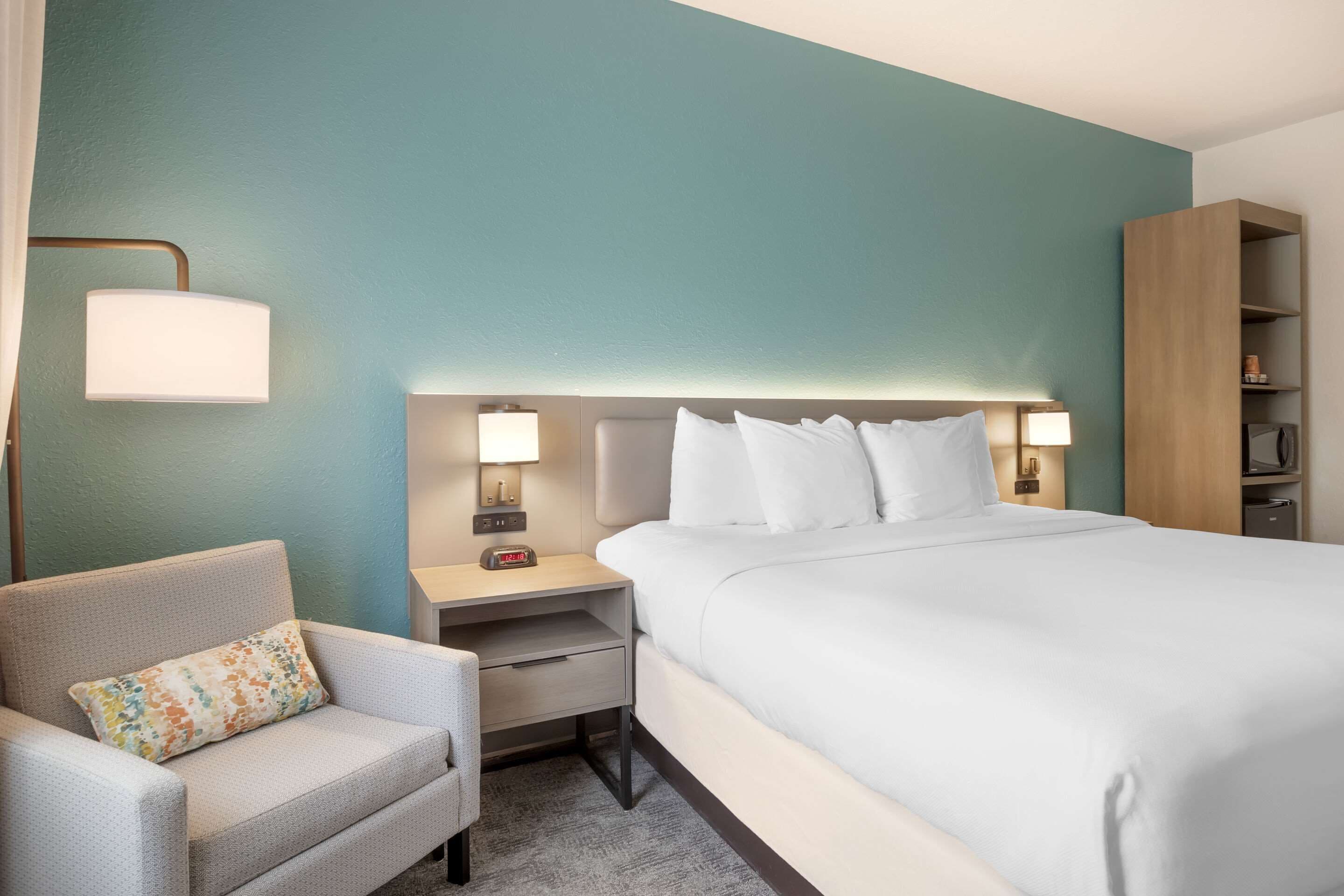 A hotel room with a bed, a chair, a nightstand with a lamp, and a wooden wardrobe.