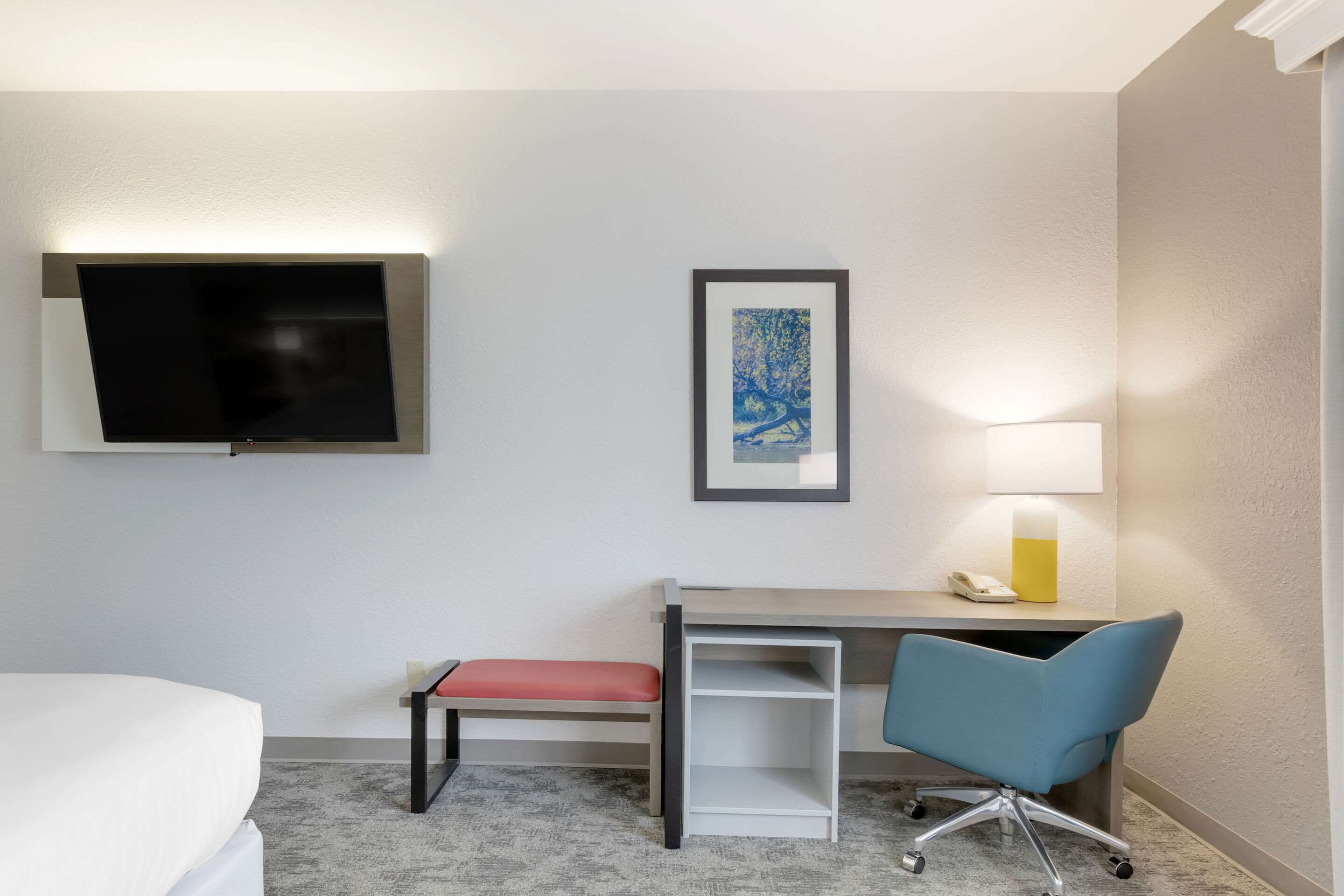 Habitación de hotel con cama, escritorio con silla, televisor en la pared, un cuadro enmarcado y lámpara de noche.