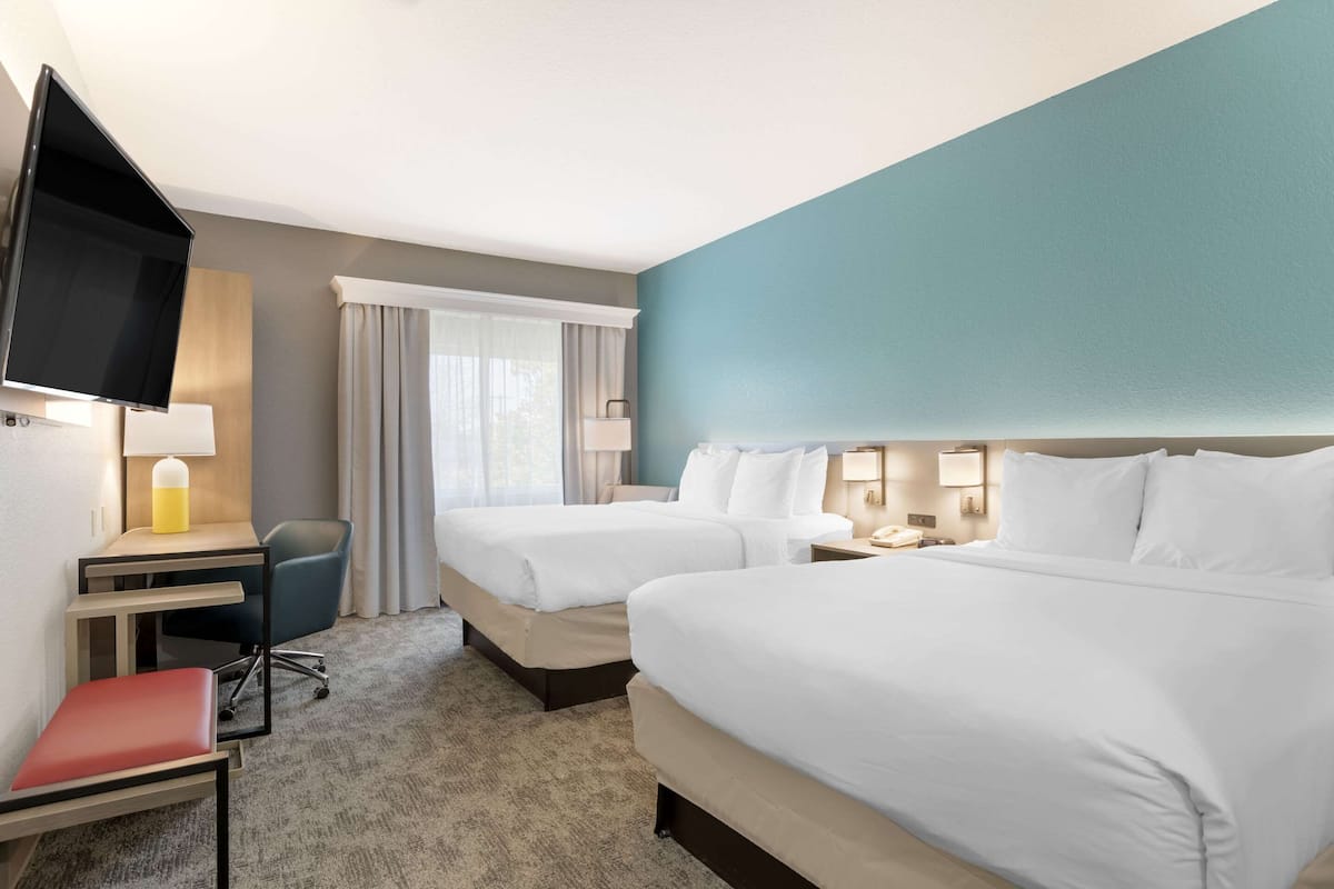Habitación de hotel con dos camas, un escritorio con silla, televisor montado en la pared y ventana con cortinas.