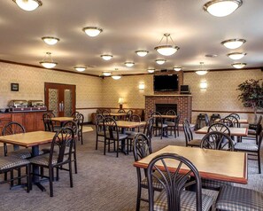 A hotel dining area with multiple tables and chairs, a fireplace, and a television mounted above it.