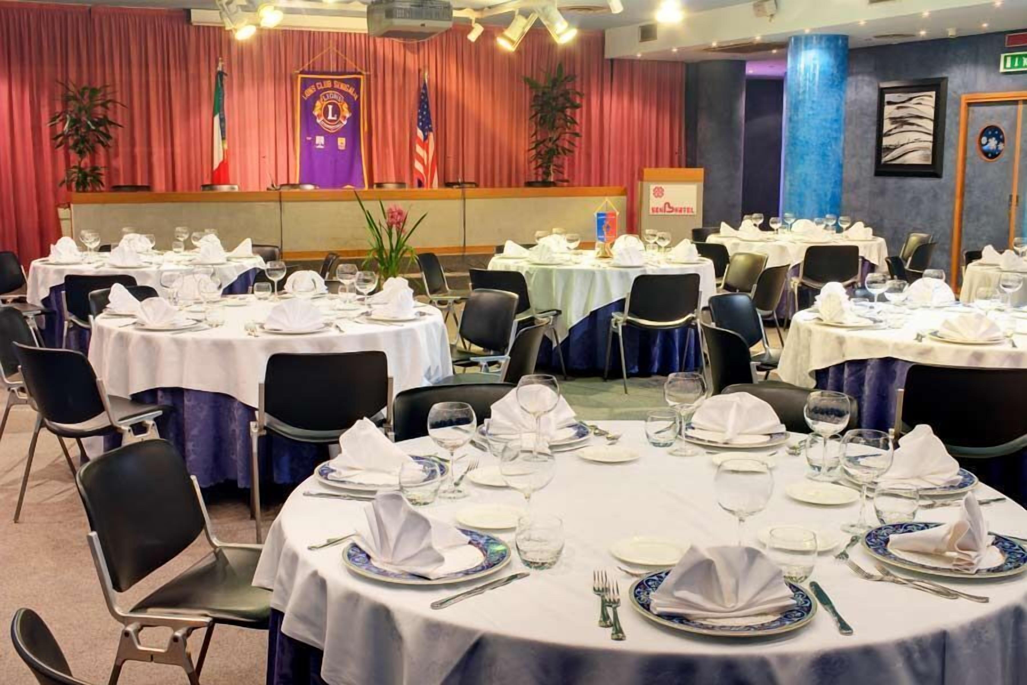 A banquet hall with round tables set for dining, chairs, and a stage with flags and banners.