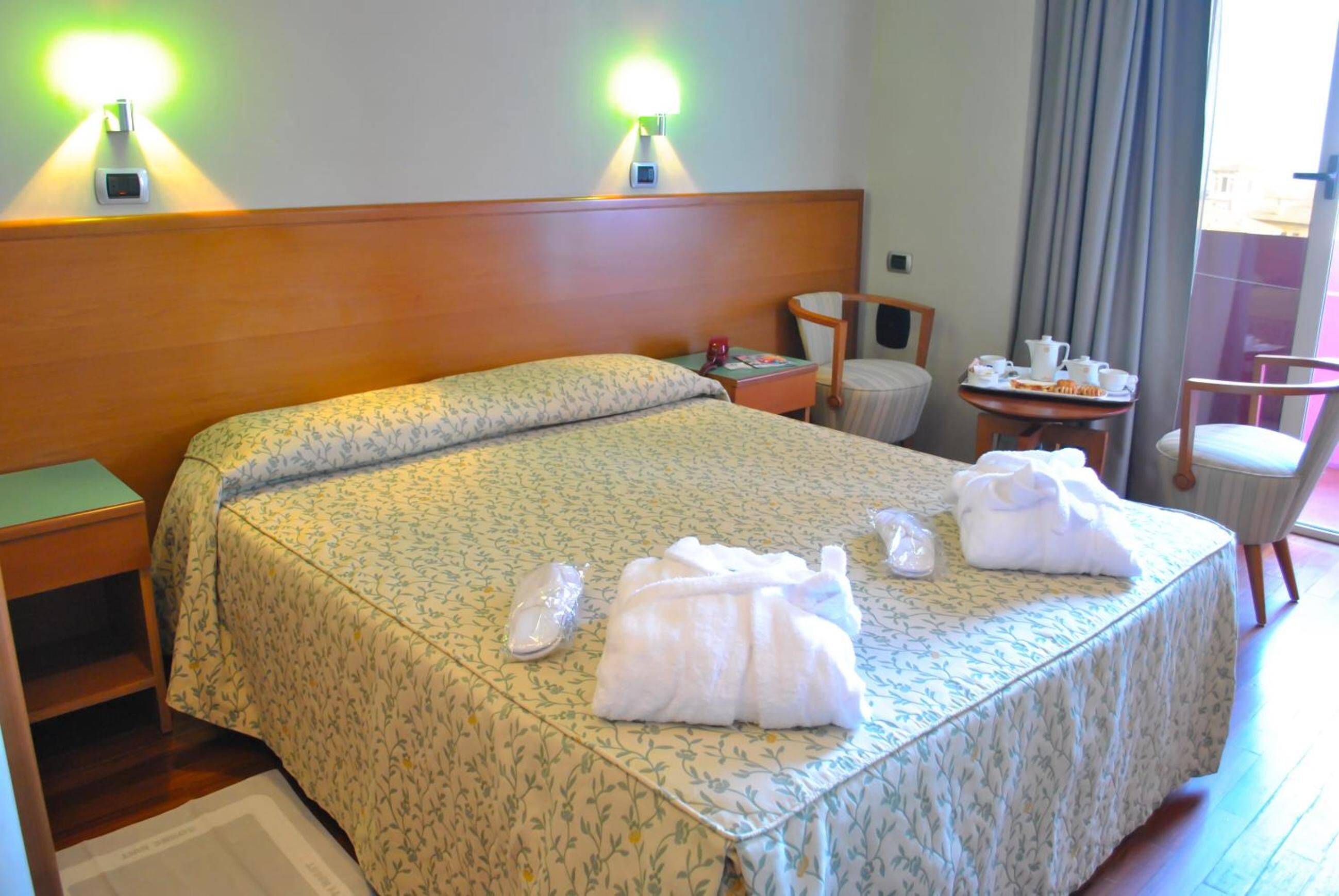 A hotel room with a large bed, two bedside lamps, a small table with breakfast items, and a chair.