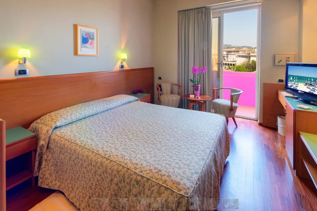 Habitación de hotel con una cama grande, dos lámparas de noche, un televisor, una mesita con un jarrón de flores y vistas a la ciudad a través de una puerta al balcón.