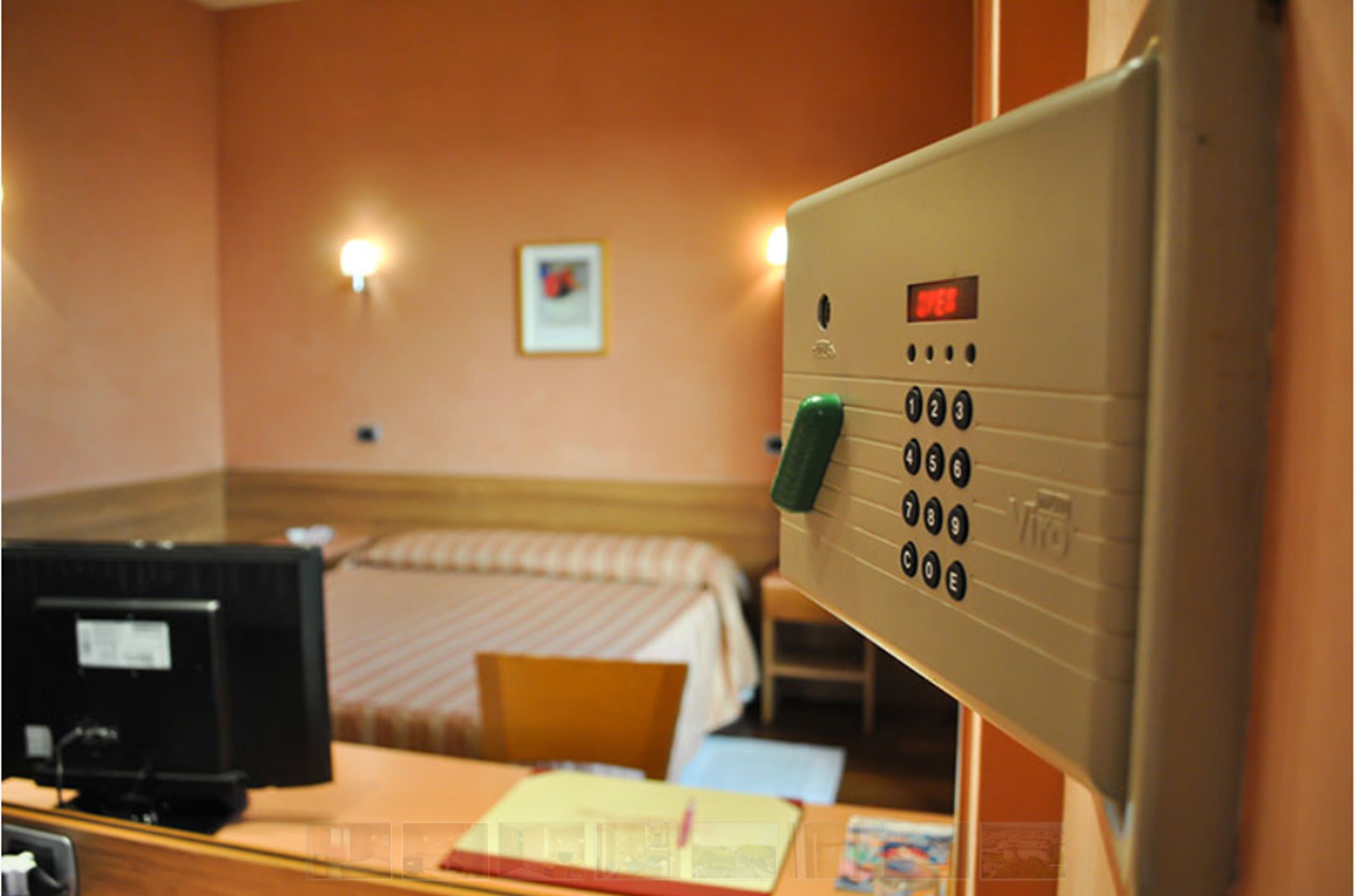 A hotel room with a bed, a desk with a computer, a telephone, and a control panel with a digital display.