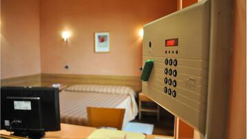 A hotel room with a bed, a desk with a computer, a telephone, and a control panel with a digital display.