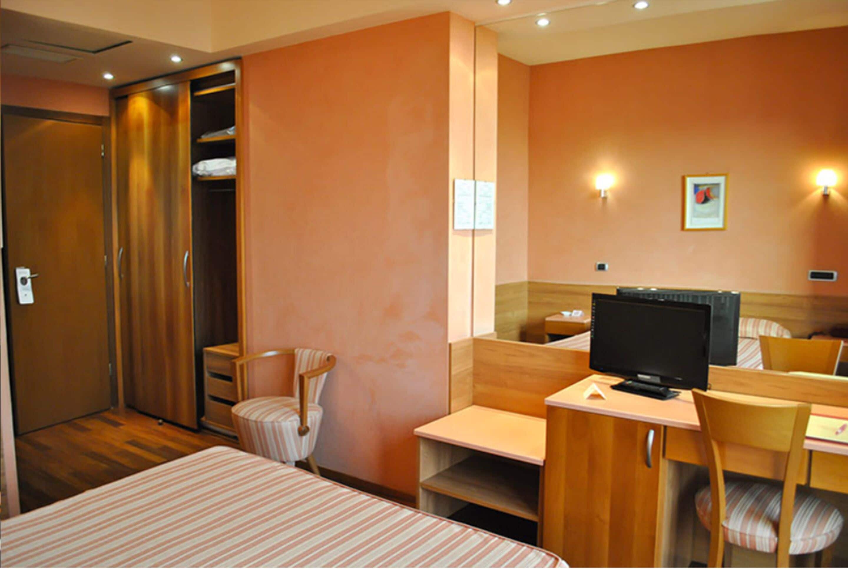 Una camera d'albergo con un letto, una scrivania con un computer, una sedia, un armadio e uno specchio.