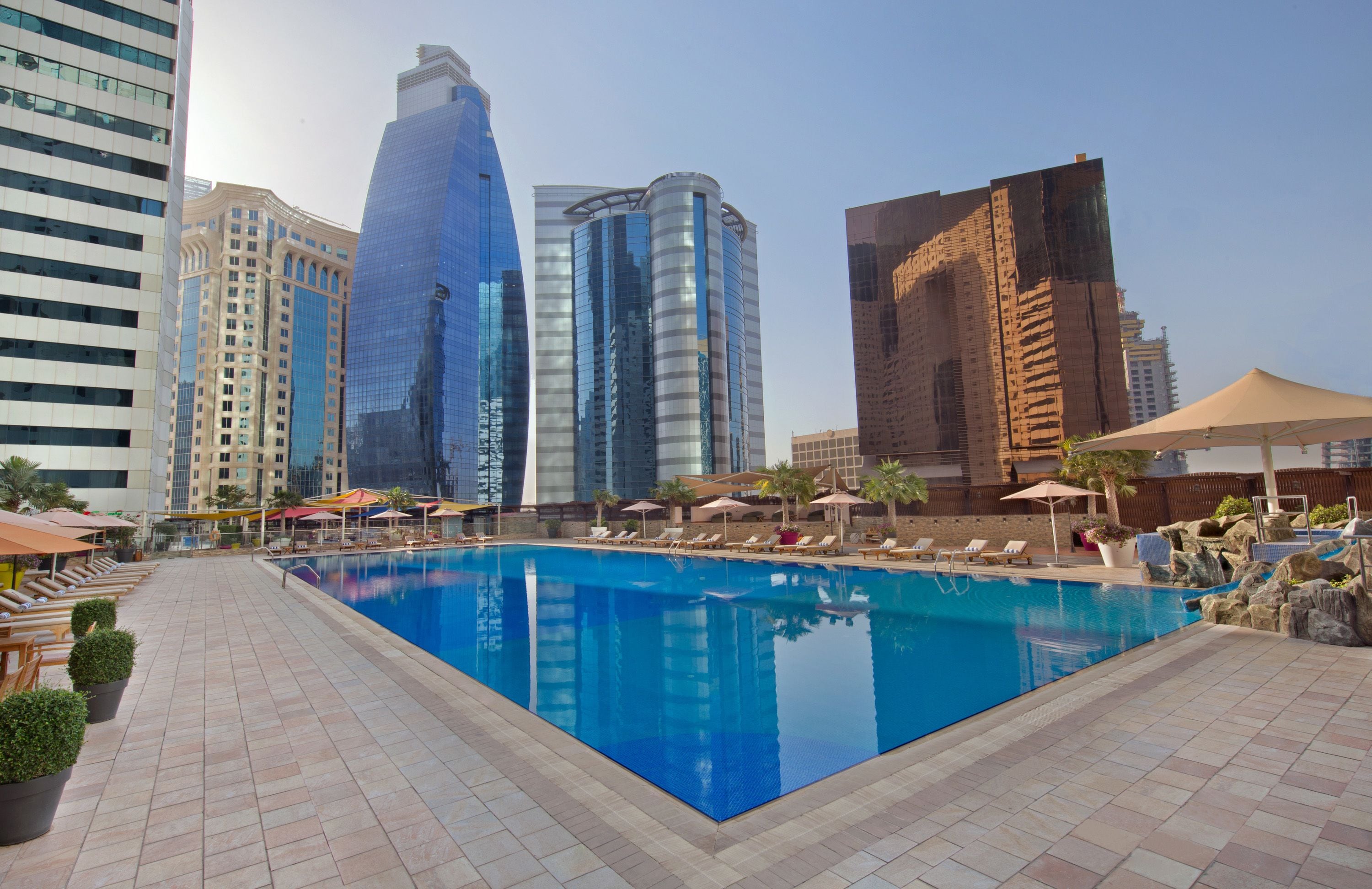 A swimming pool with lounge chairs and umbrellas, surrounded by modern buildings.