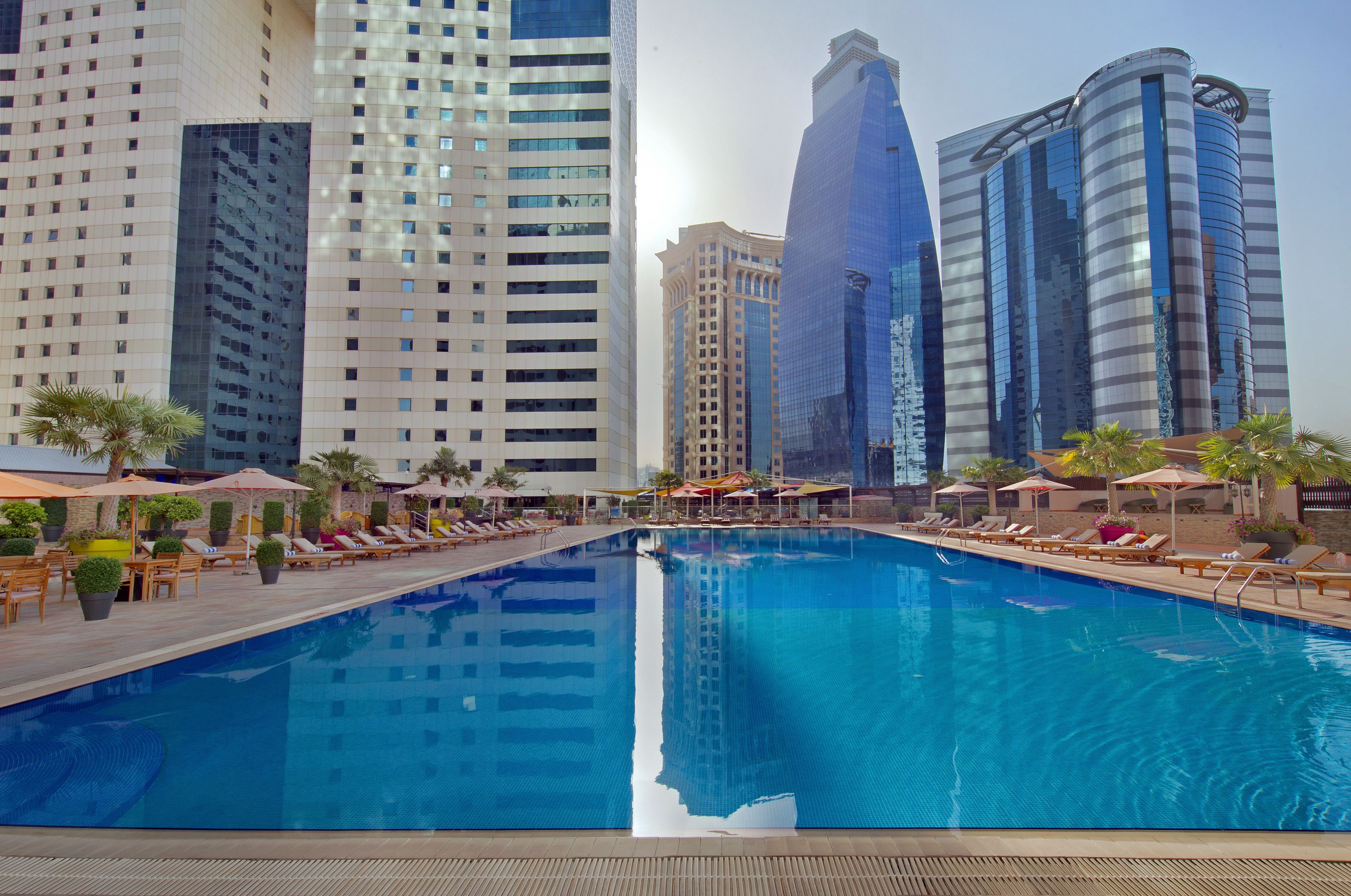 Una piscina con sillones de descanso y sombrillas, rodeada de edificios altos.