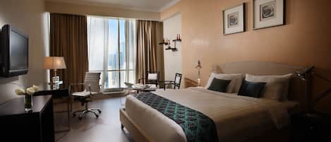 Habitación de hotel con una cama grande, un escritorio con televisor, una silla, una mesita y una ventana con cortinas.
