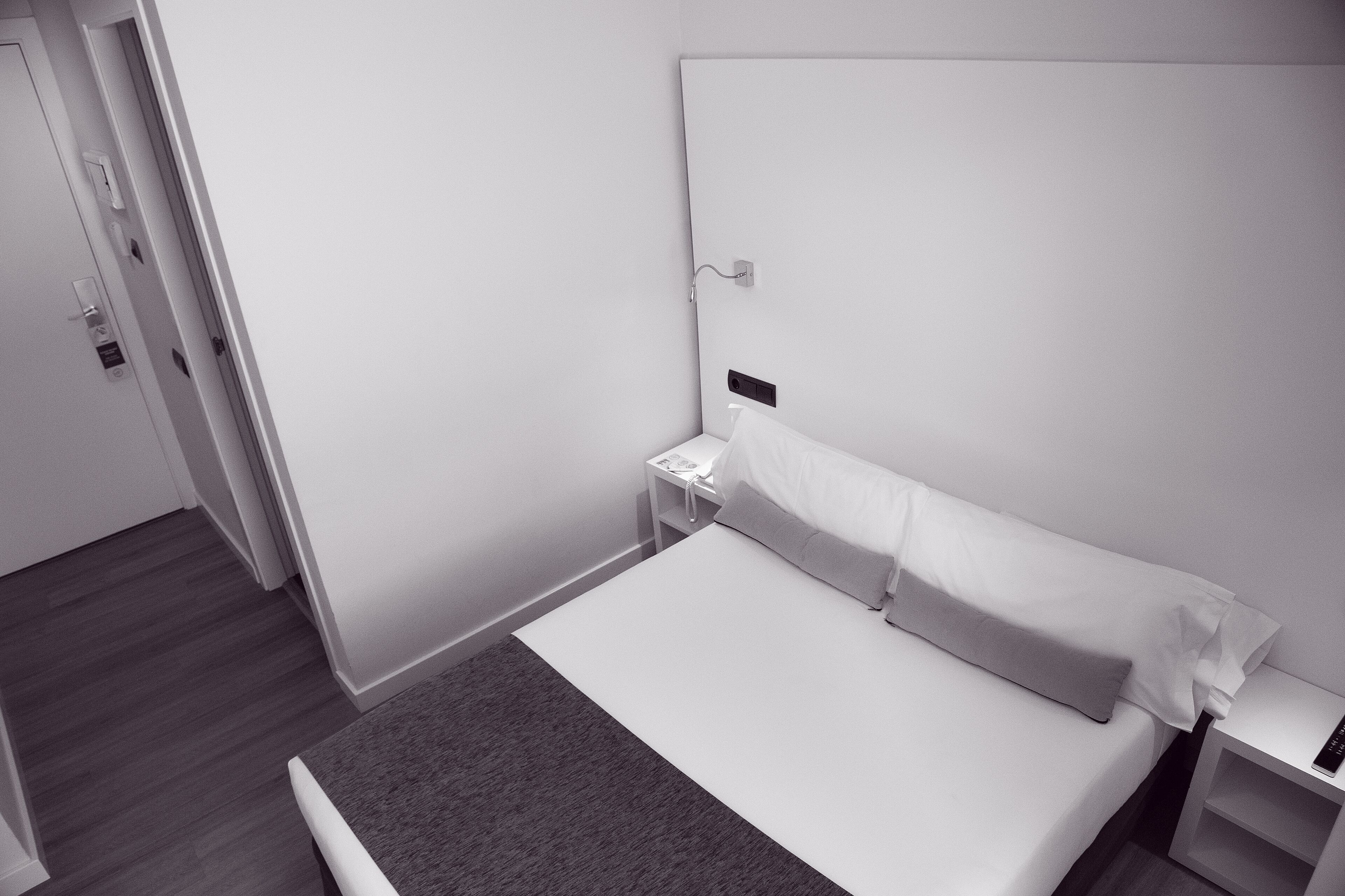 A modern hotel room with a neatly made bed, a bedside table with a lamp, and a door leading to another room.