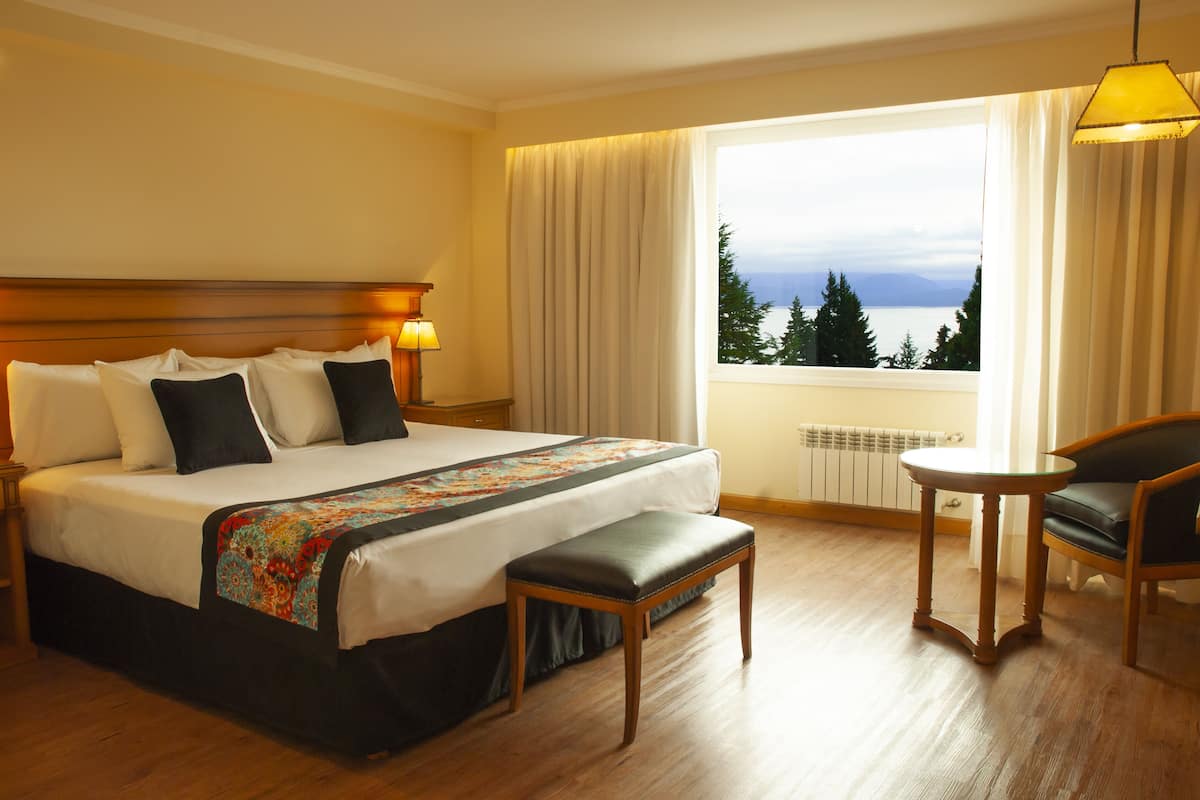 A hotel room with a large bed, a bench, a small table, and a view of the outdoors.