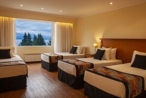 A hotel room with four single beds, a large window with curtains, a wooden floor, and a nightstand with a lamp.