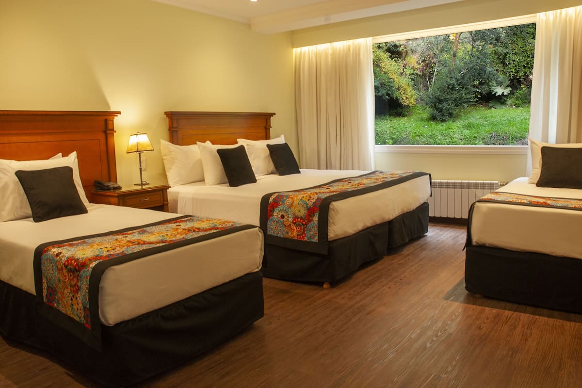 A hotel room with three beds, a nightstand with a lamp, a window with curtains, and a view of greenery outside.