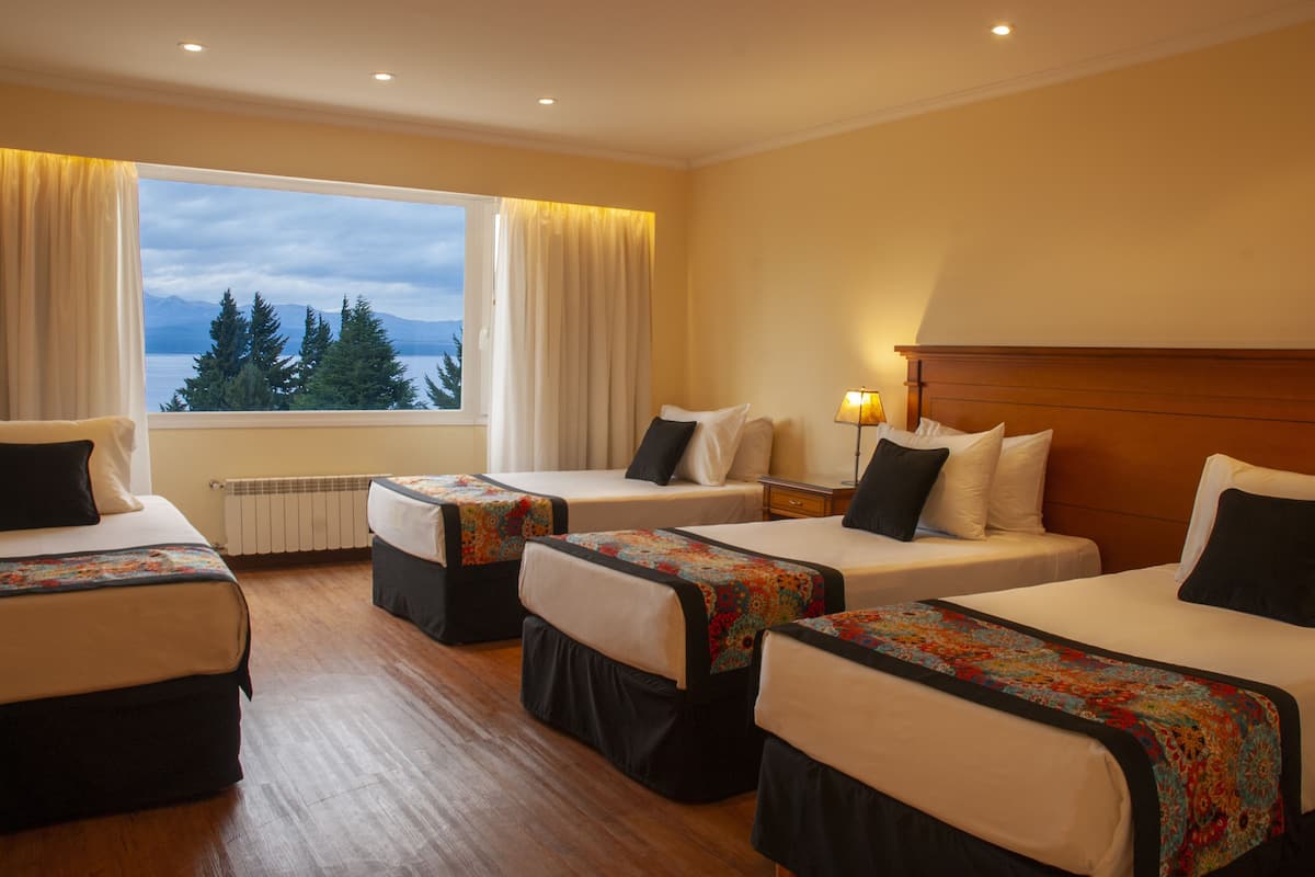 A hotel room with four beds, a large window with a view of trees and mountains, a wooden floor, and a nightstand with a lamp.