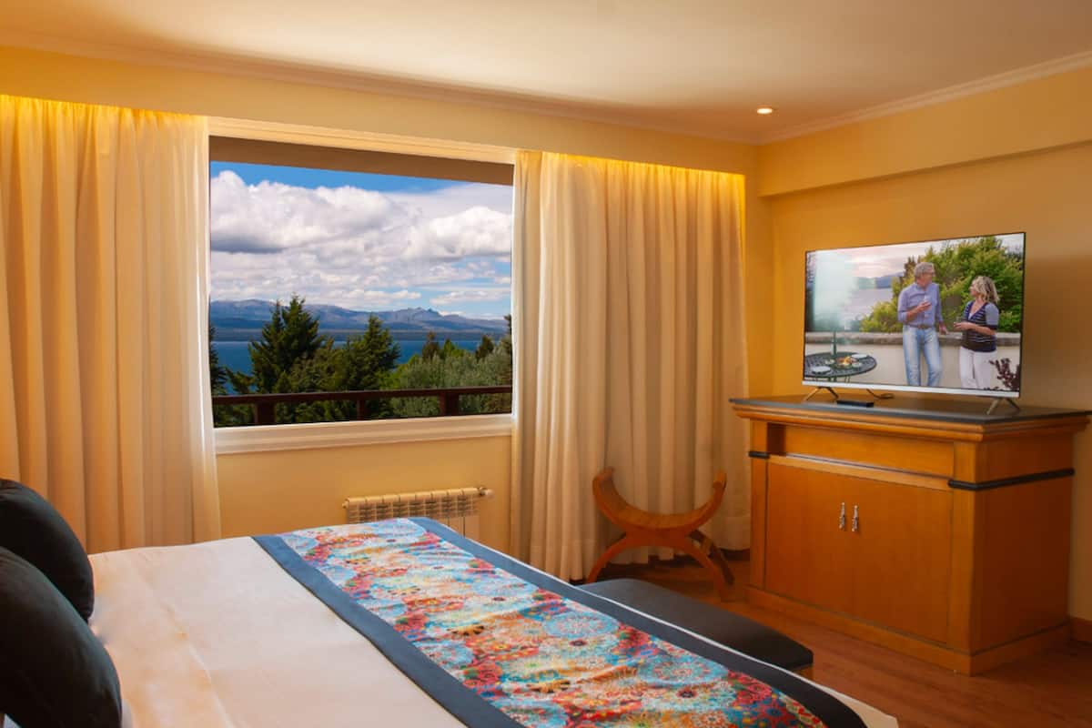 A hotel room with a bed, a TV, a wooden cabinet, and a window with a mountain view.