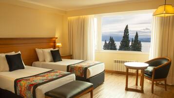 Quarto de hotel com duas camas, uma mesa pequena, uma cadeira e vista para árvores e montanhas pela janela.