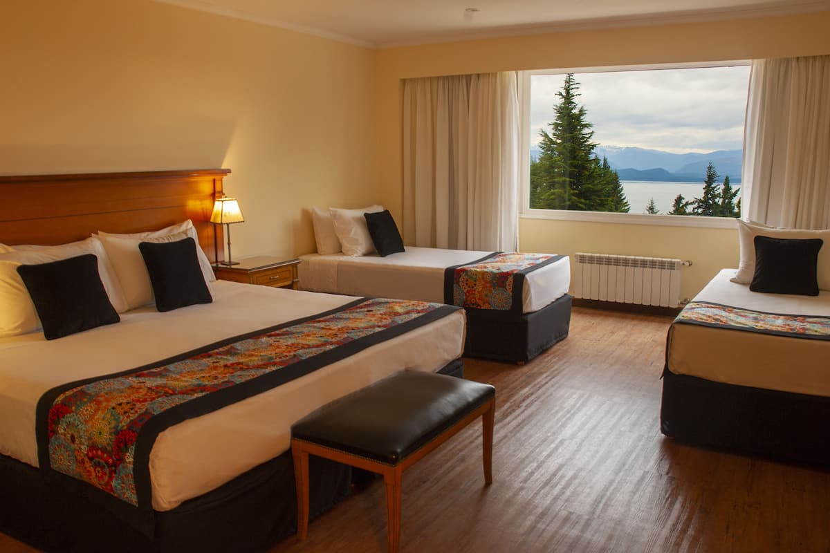A hotel room with two beds, a bench, a window with a view of mountains and a lake, and a radiator.