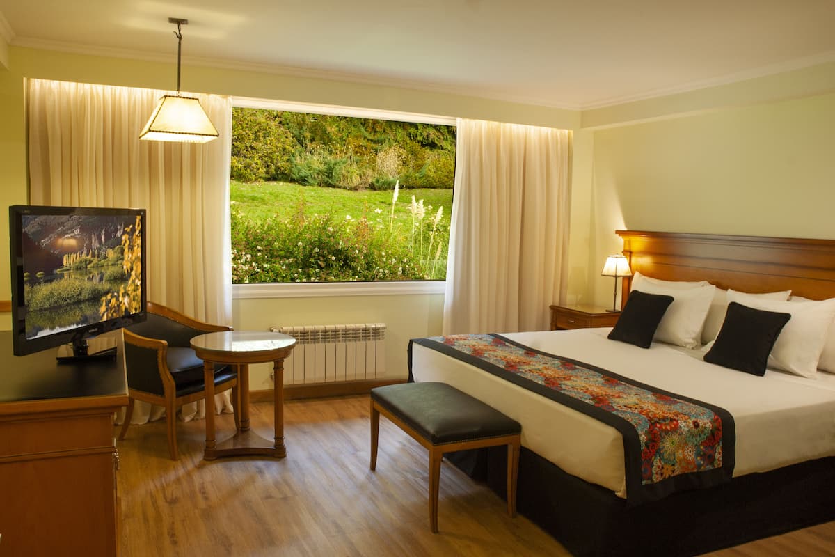 A hotel room with a bed, a desk with a television, a chair, a window with curtains, and a view of a garden.