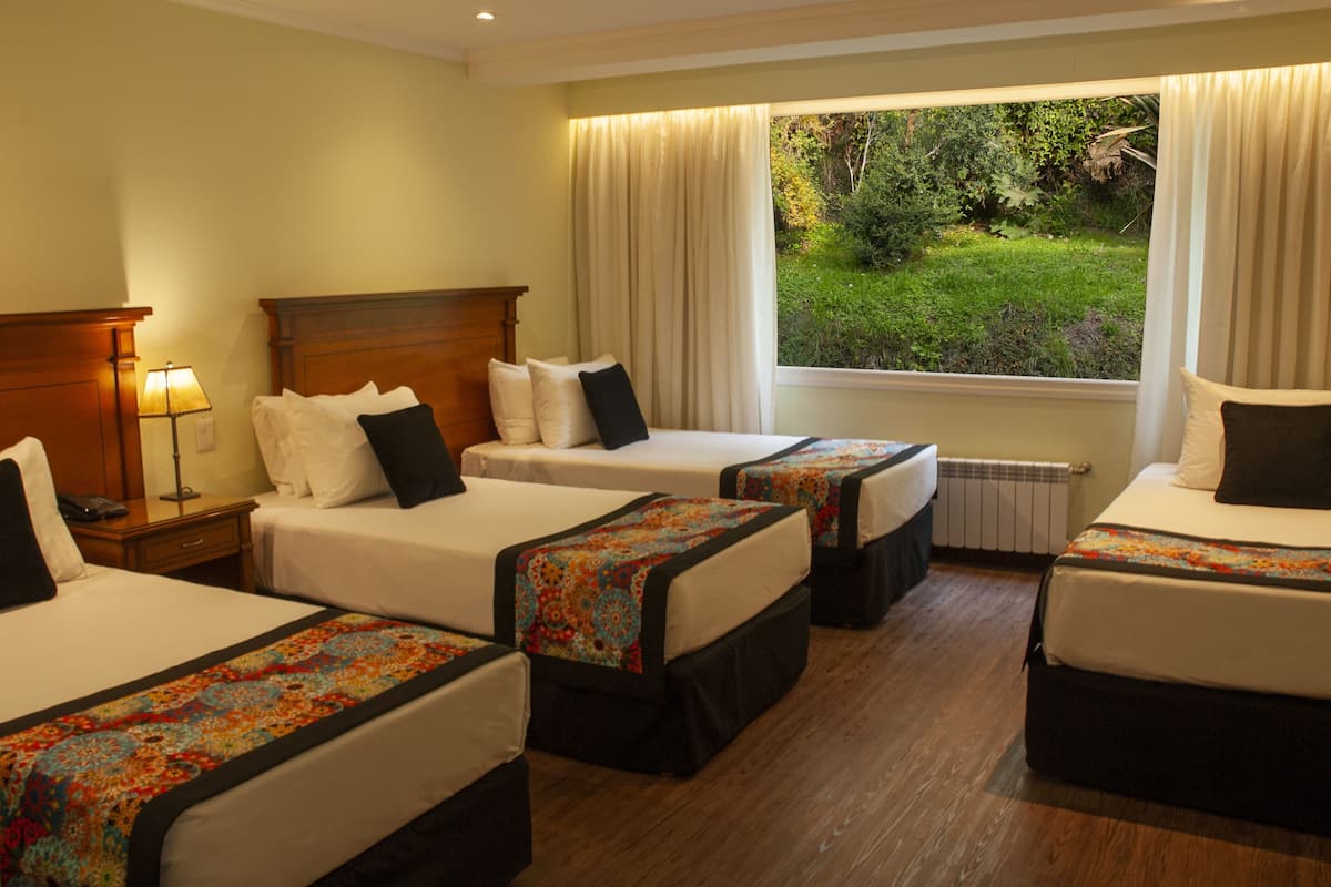A hotel room with three beds, a wooden headboard, a window with curtains, and a view of greenery outside.