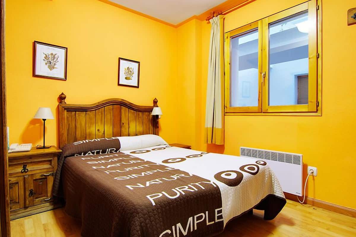 A room with a bed, wooden headboard, two bedside tables with lamps, a window with curtains, and a wall-mounted air conditioner.