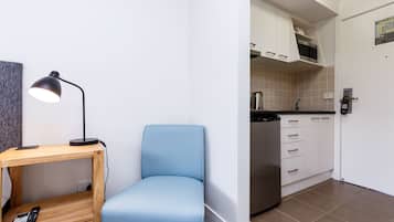 A room with a blue chair, a wooden table with a lamp, and a kitchen area with white cabinets and a stainless steel appliance.