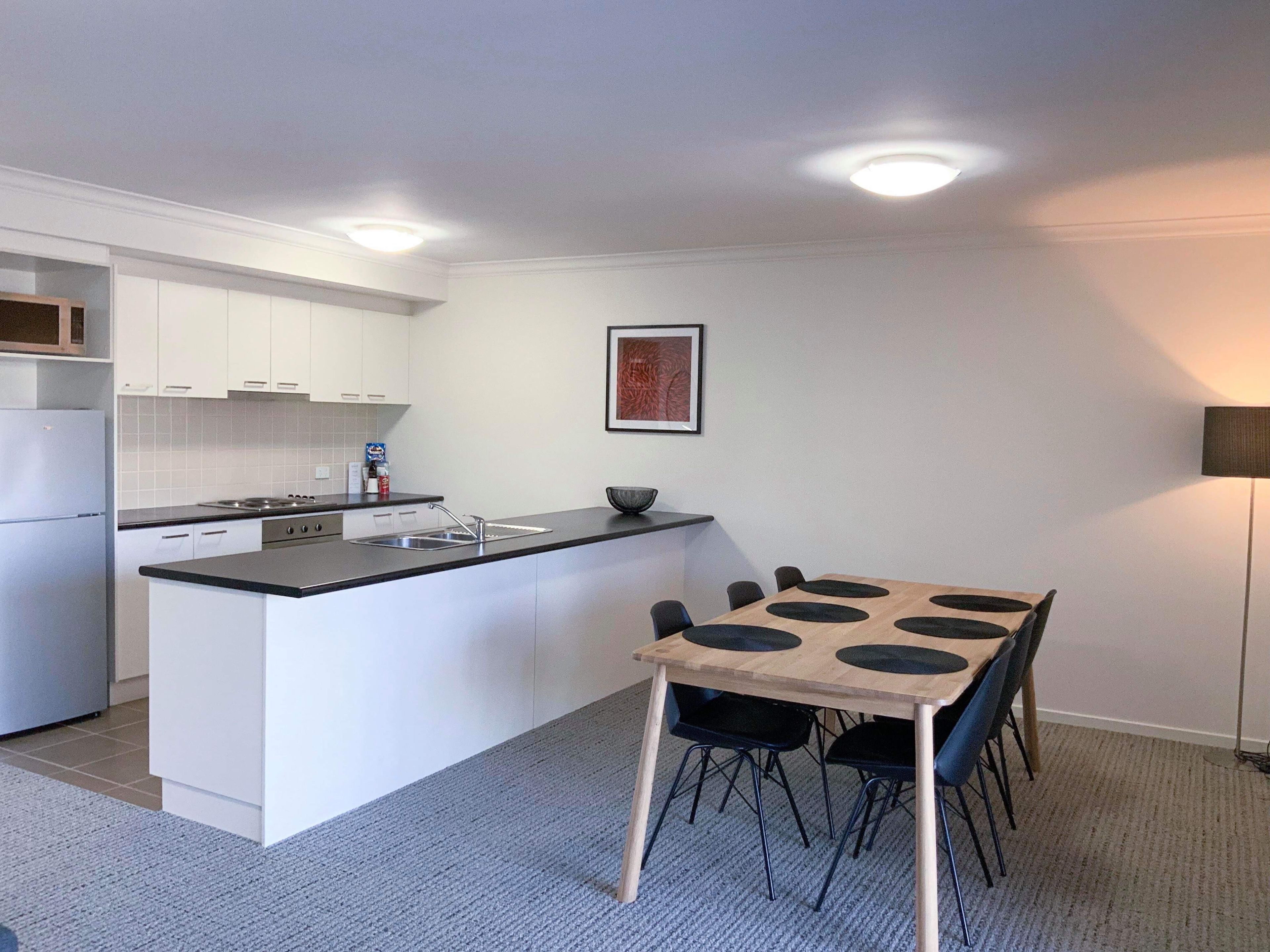 A compact kitchen with white cabinets, a black countertop, and a central island with a sink. There is a dining area with a table and chairs, and a floor lamp.