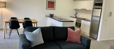 A modern living room with a dark sofa, a wooden coffee table, and a dining area with a table and chairs. A kitchen with white cabinets and stainless steel appliances is visible in the background.