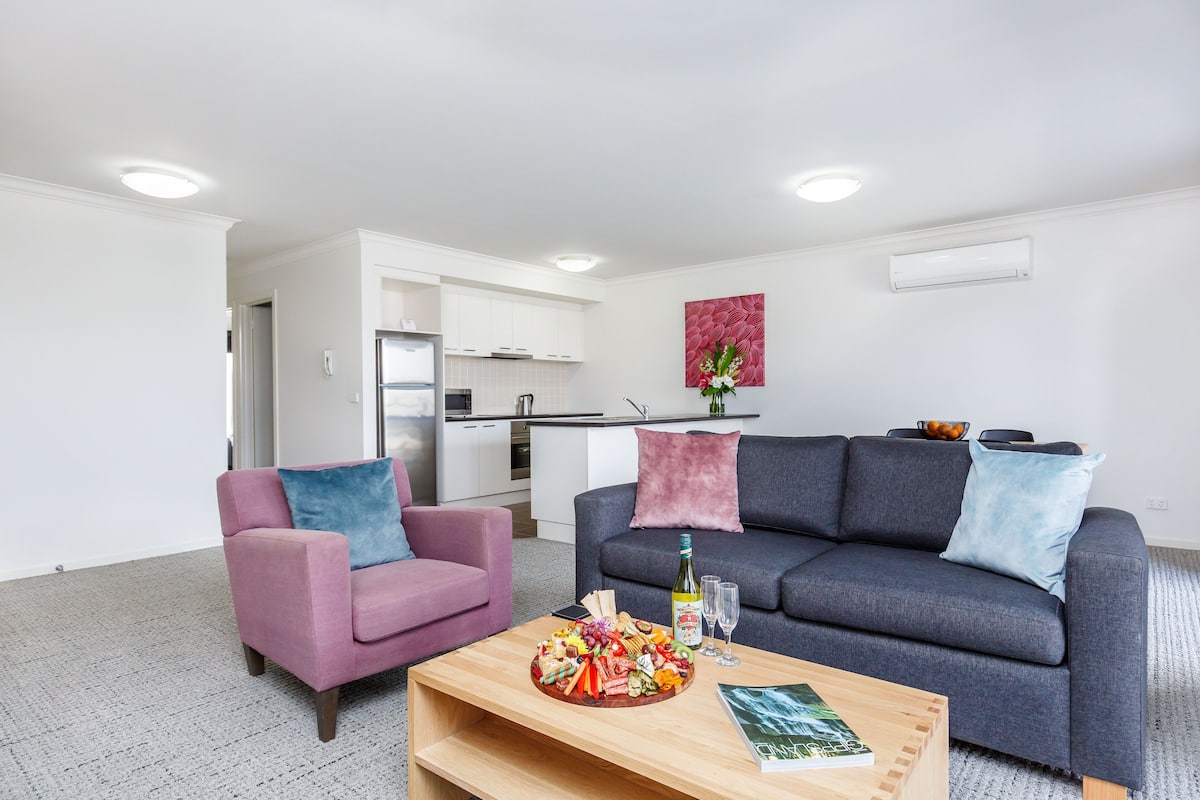 Una sala moderna con un sillón rosa, un sofá gris y una mesa de centro de madera con aperitivos y bebidas.