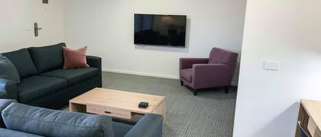 A living room with a dark green sofa, a light wooden coffee table, a purple armchair, and a flat-screen TV mounted on the wall.