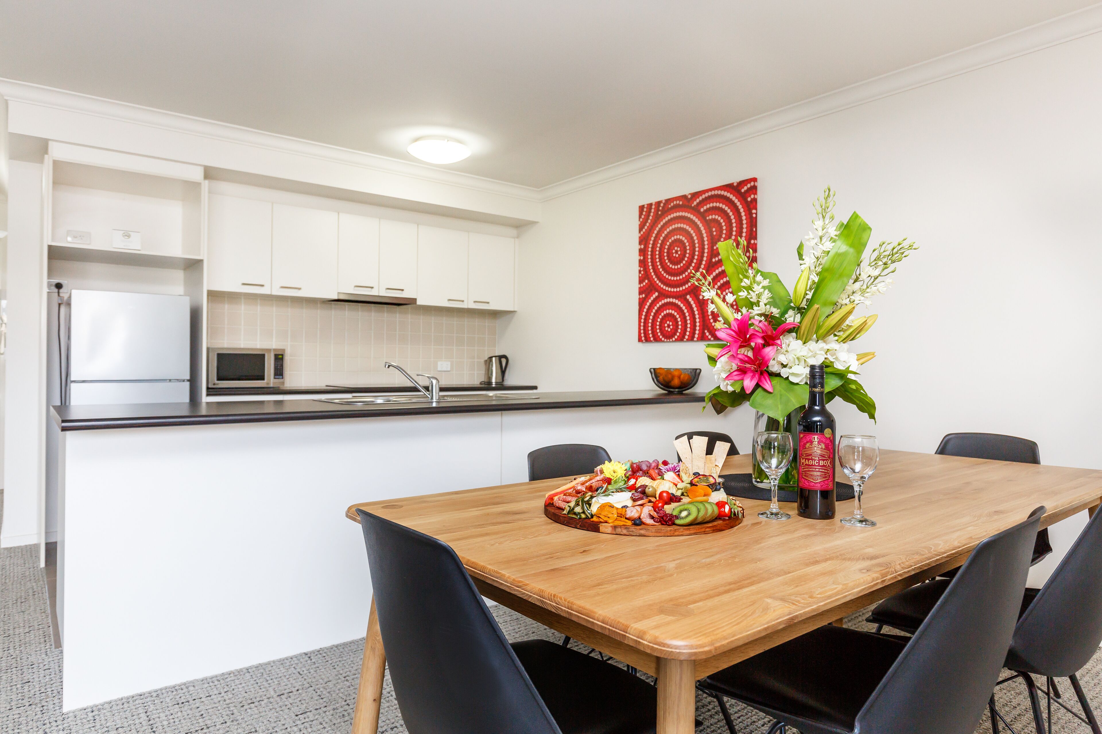 A modern kitchen with white cabinets, a microwave, and a sink. A wooden dining table with chairs is set with a fruit platter and wine glasses.
