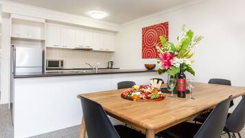 A modern kitchen with white cabinets, a microwave, and a sink. A wooden dining table with chairs is set with a fruit platter and wine glasses.